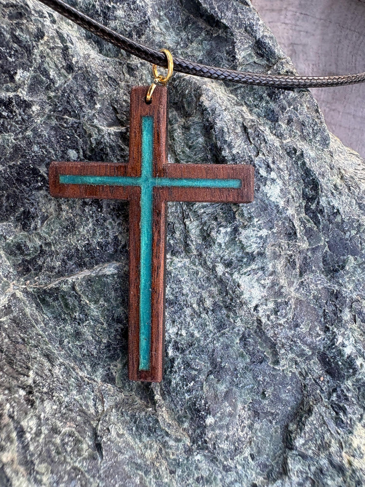 Black walnut cross necklace with turquoise resin inlay and black cord on dark rock