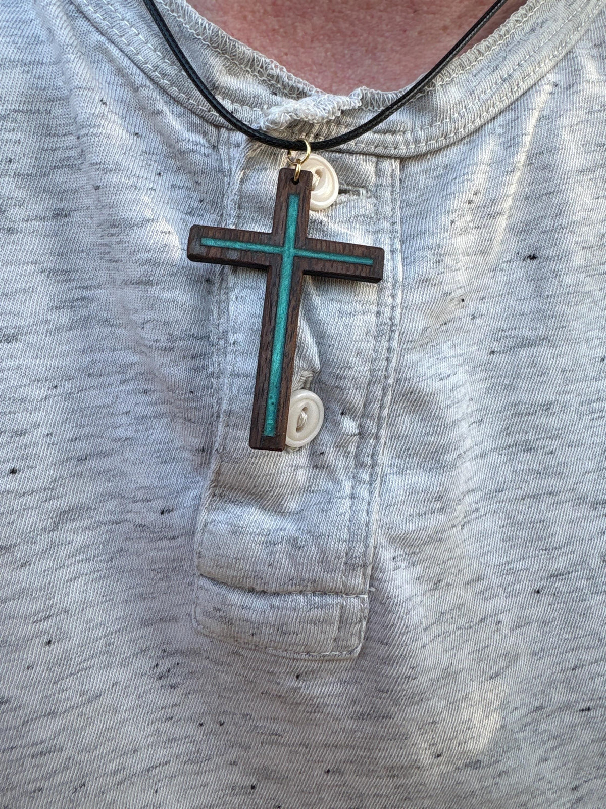 Turquoise resin-inlay black walnut cross necklace on a black cord worn over a light gray shirt