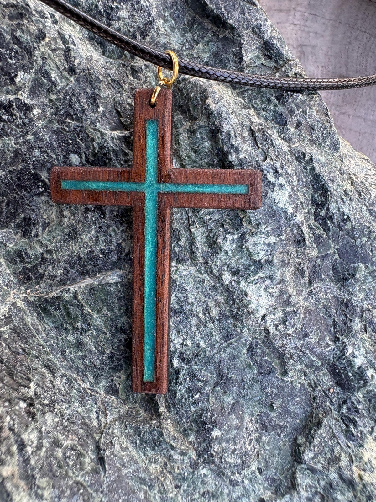 Black walnut cross necklace with turquoise resin inlay and black cord on dark rock