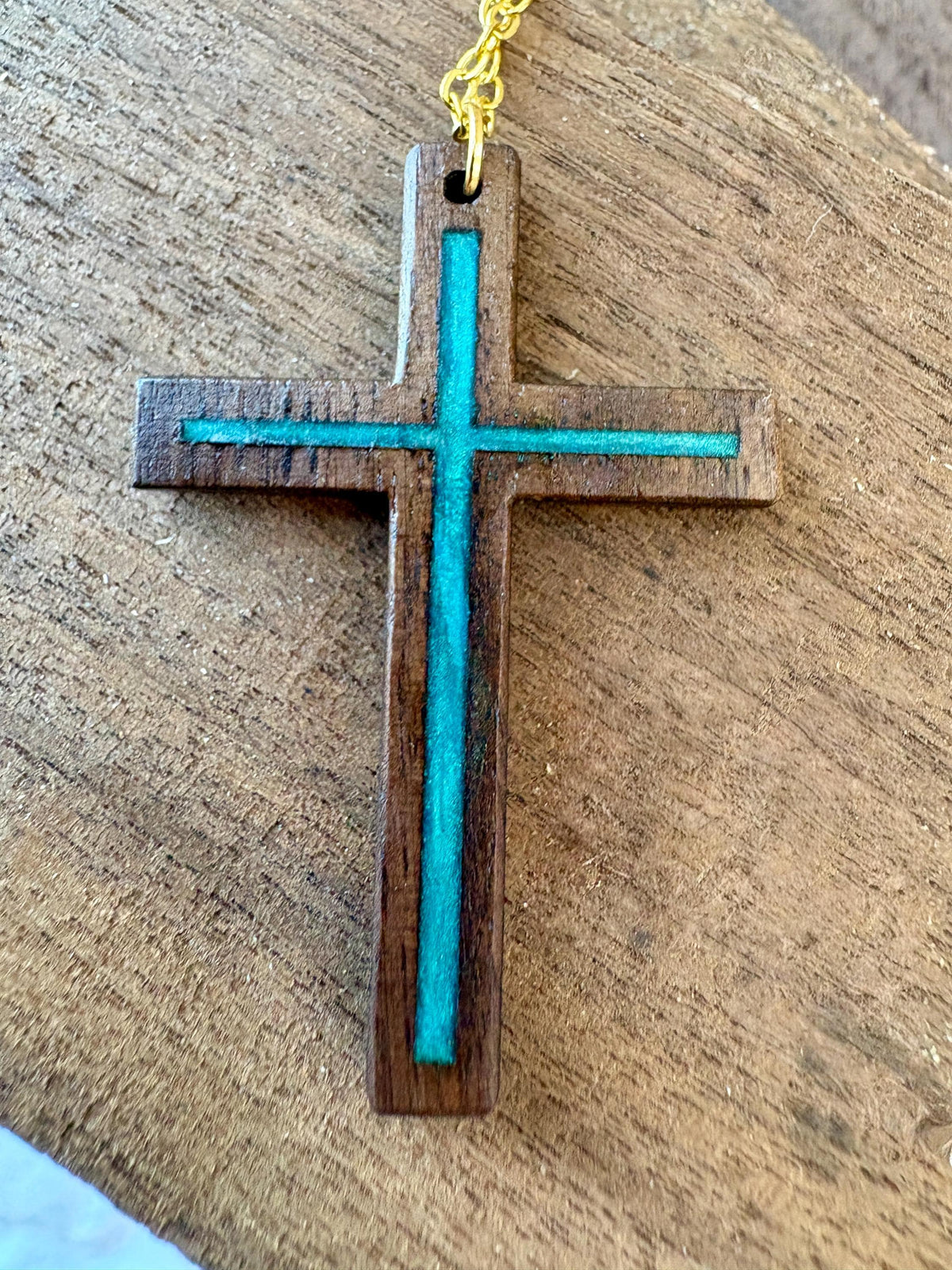 Close-up of black walnut cross pendant with turquoise resin inlay and gold-colored chain on wood