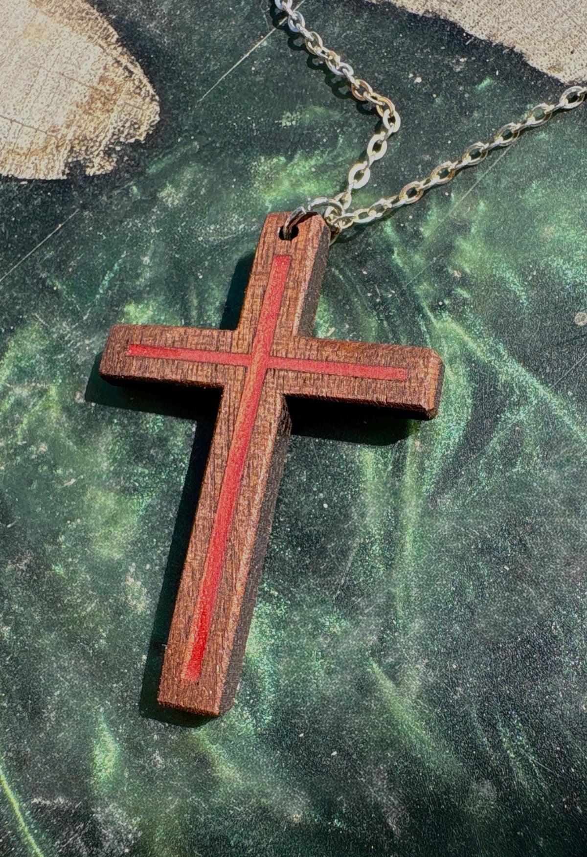 Black walnut cross pendant with red resin inlay displayed on a green resin-and-wood surface