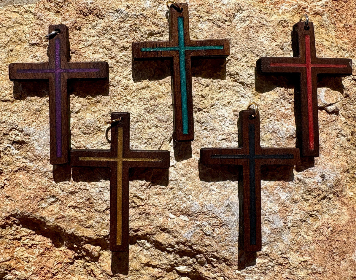 Five black walnut cross pendants with colorful resin inlays displayed on textured tan rock
