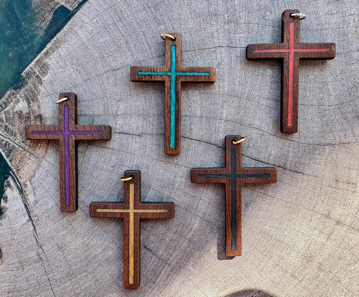 Five black walnut cross pendants with purple, turquoise, red, gold, and blue resin inlays arranged on gray wood