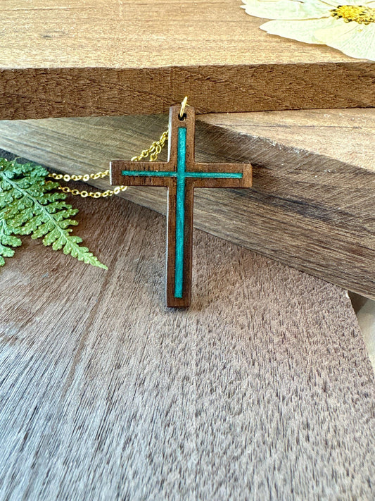Black walnut cross necklace with turquoise resin inlay and gold-colored chain against stacked wood beside a fern