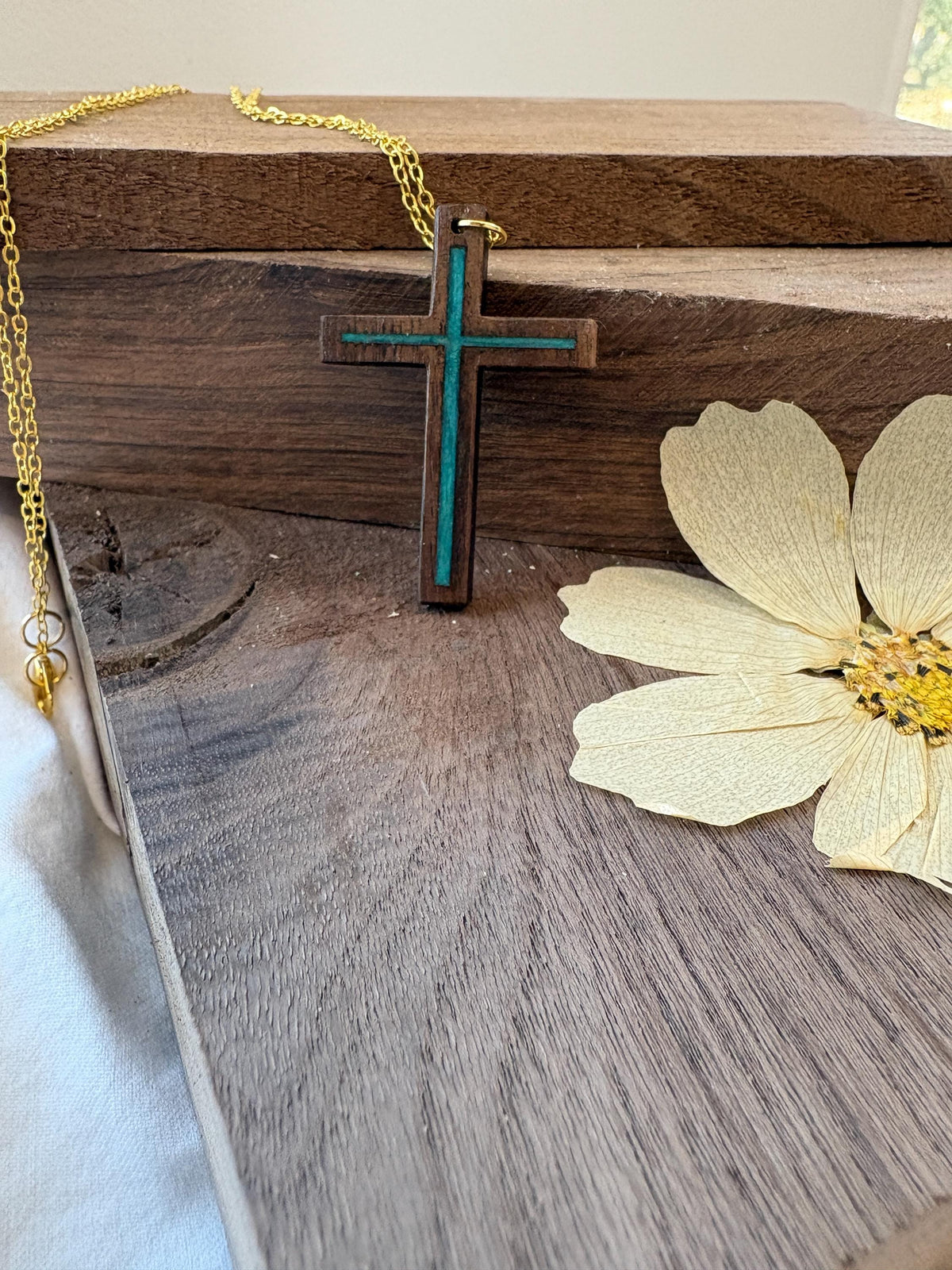 Turquoise resin-inlay walnut cross necklace with gold-colored chain displayed on wood beside a pale flower